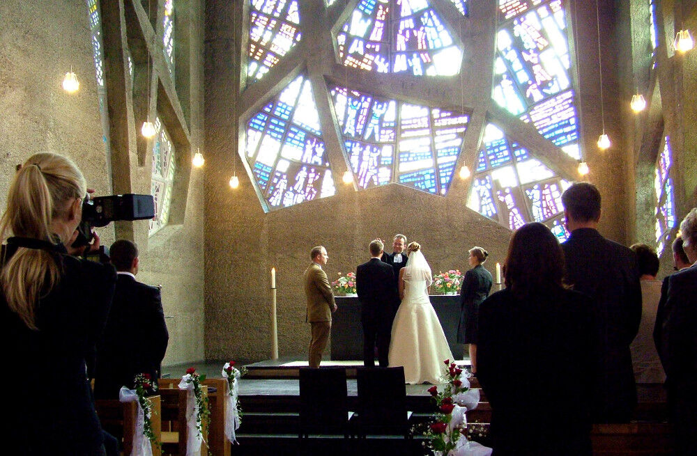 Innenraum einer Kirche mit bunten Fenstern, wo ein Brautpaar vor dem Altar steht